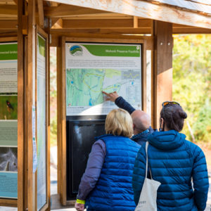 Three visitors stand in front of a large trailhead kiosk, one points to a map on the kiosk.
