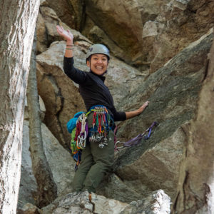 A climber with gear on her harness stands on the cliff, looks down and waves. 