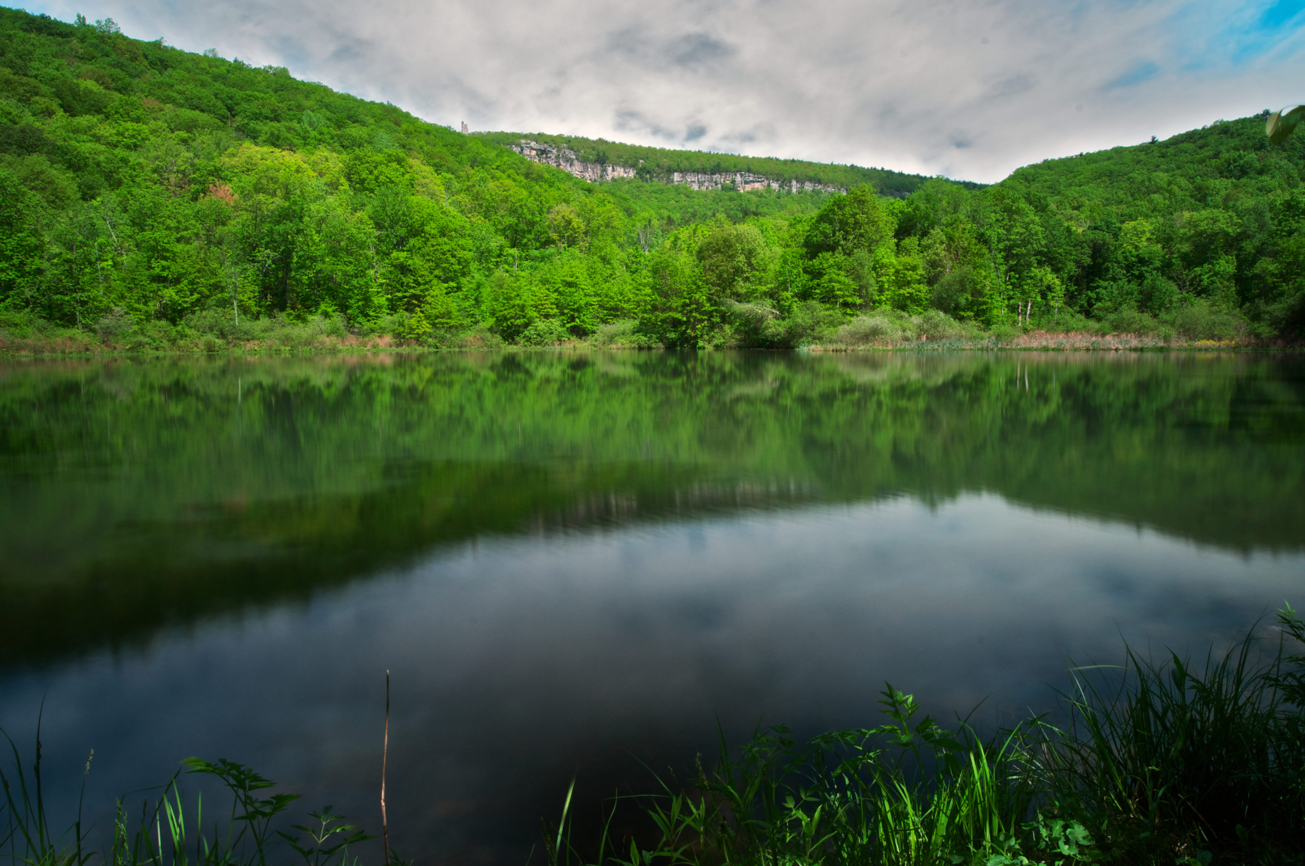 Duck Pond in the foreground, reflecting the background of Skytop tower and cliffs against a cloudy sky and forested hills rise between