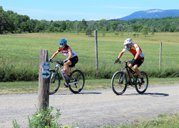 Two people ride bicycles on Lenape Lane, a gravel road, with a field behind them and the cliffs of Millbrook in the distance.