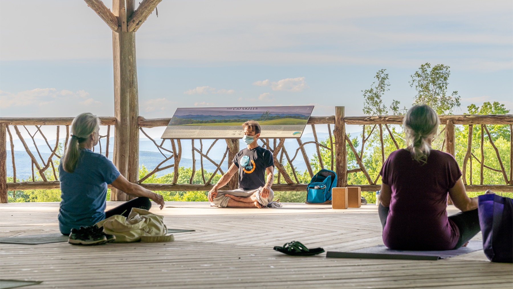 A yoga instructor wearing a mask faces his class in front of the railing at the Slingerland Pavilion. Two students face him
