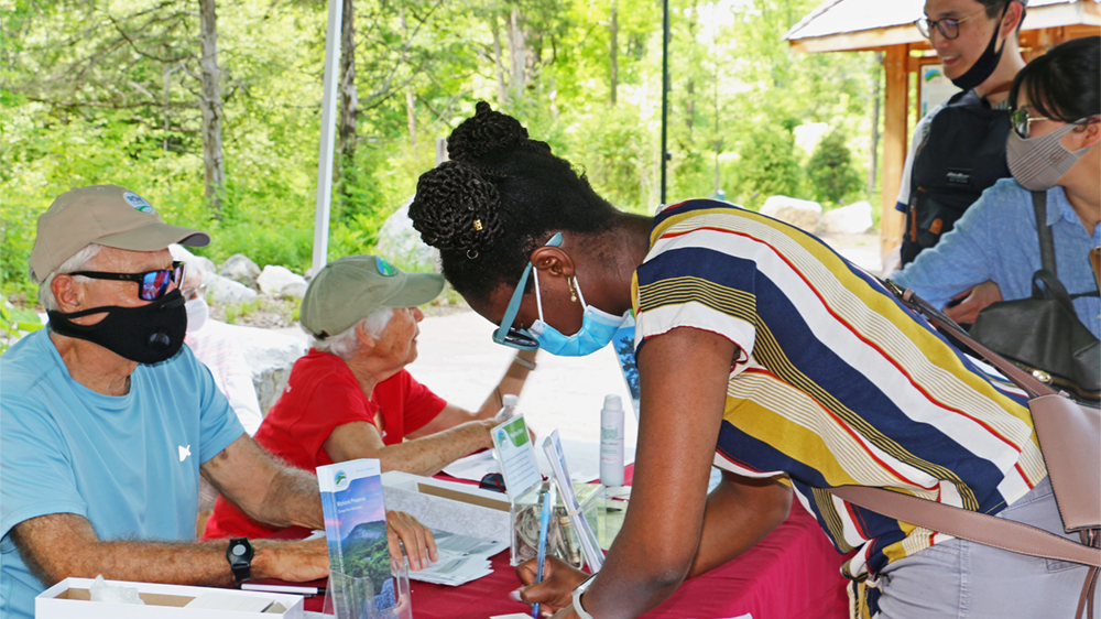 A visitor leans on a table to sign up for a Healthy Ulster pass. Sitting behind the table, a volunteer looks on.