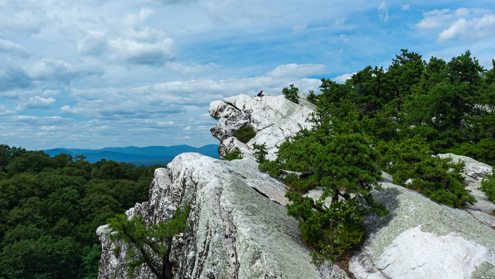 A vista featuring the top of Bonticou Crag with the Catskill mountains in the background. A hiker sits on top of the crag.