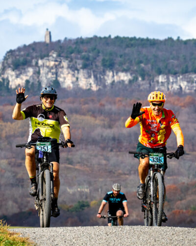 Two bike riders waving as they ride in the Shawangunk Grit annual gravel bike ride. Skytop tower and the Shawangunk Ridge is in the background.