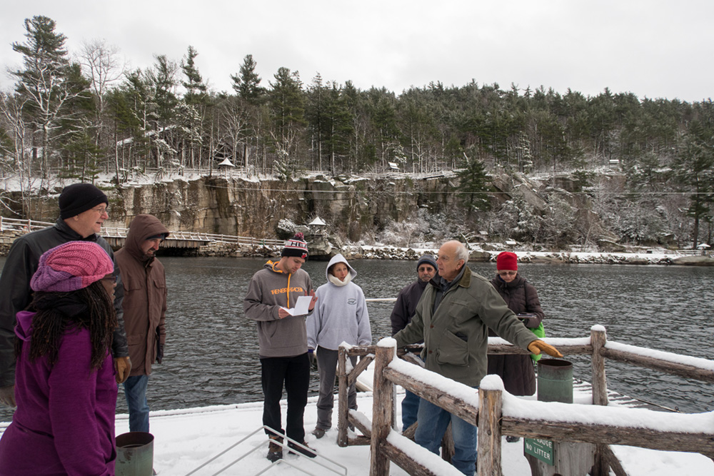 A group gathers around on the dock of Mohonk Lake for a presentation on weather recording equipment. A wintery scene of forests and cliff edges surrounding the lake are in the background.