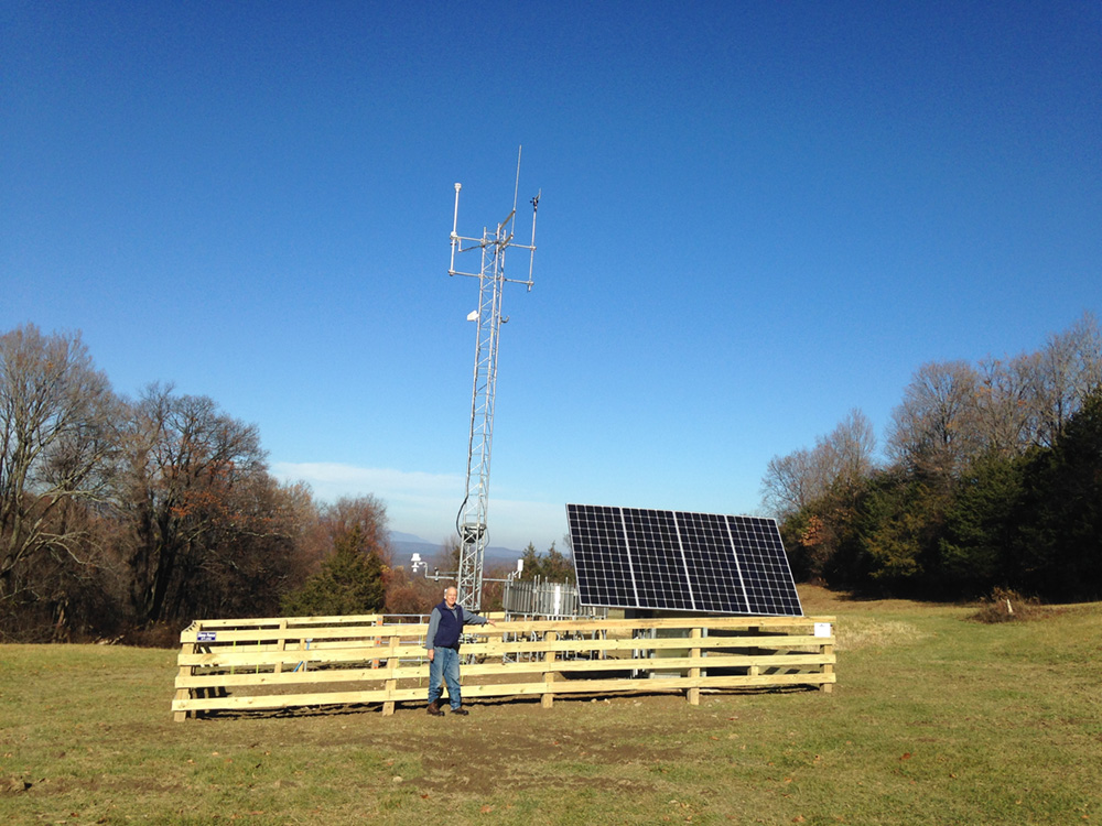 A fenced in weather station with solar panels and a large tower is in an open field with Paul Huth Director of Research Emeritus