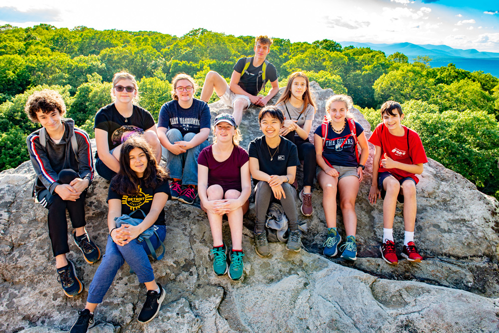 A group of teenagers sit on a cliff with a lush green forest and blue Catskill mountains in the background