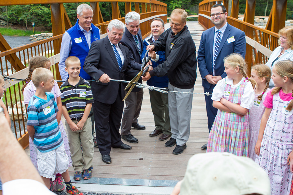 A group of adults and children gather around for the ribbon cutting ceremony for the grand opening of the Trapps bridge at Mohonk Preserve