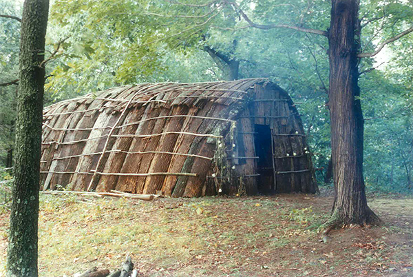 A traditional Native American longhouse constructed with durable materials sits in a forested field