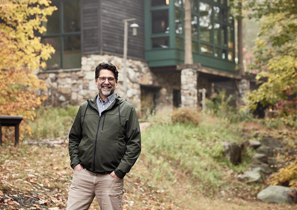 President Kevin Case stands outside the Visitor Center in a forested setting in Autumn