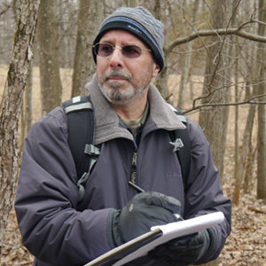 Ralph Durham in a wooded outdoor area with a clipboard taking field study notes
