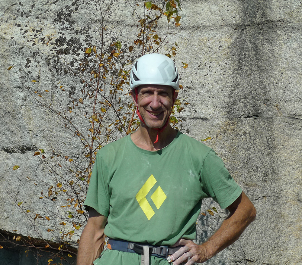 Chair Russ Clune stands in front of a cliff with a rock climbing helmet and harness on.