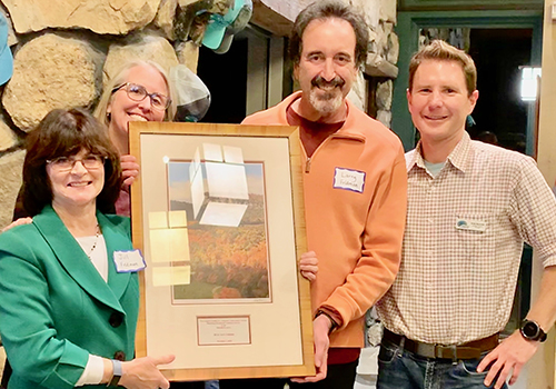 Volunteer manager Andy Reynolds poses with three other individuals with a framed photo of Skytop Tower in the Mohonk Preserve Visitor Center