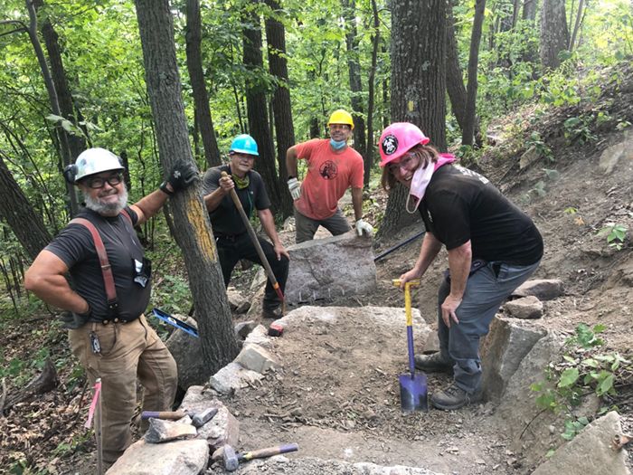 Four trail builders wearing hard hats while posing for a photo over a trail building project.