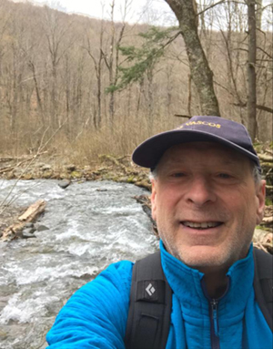 Hiker smiling in foreground with a forest and stream in background