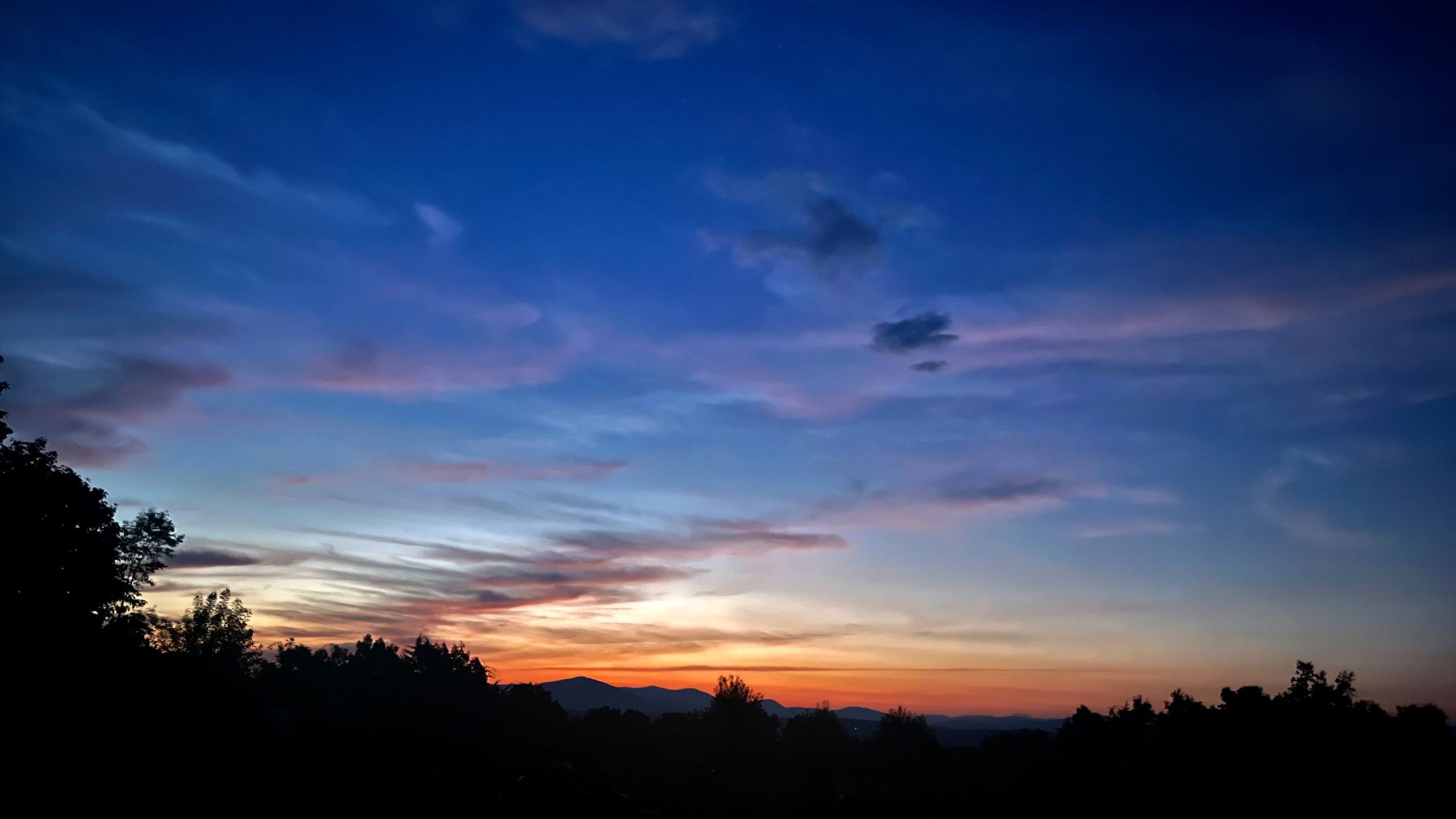 Melissa_Styles_image00001 Colorful streaks of blue, purple, and orange fill the evening sky above distant hills, with silhouetted trees framing the sunset landscape at Mohonk Preserve.