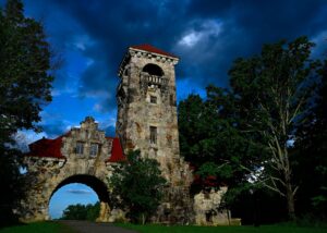 The stone Testimonial Gateway stands beneath dramatic dark storm clouds, with sunlight illuminating sections of the historic structure and surrounding trees at Mohonk Preserve.